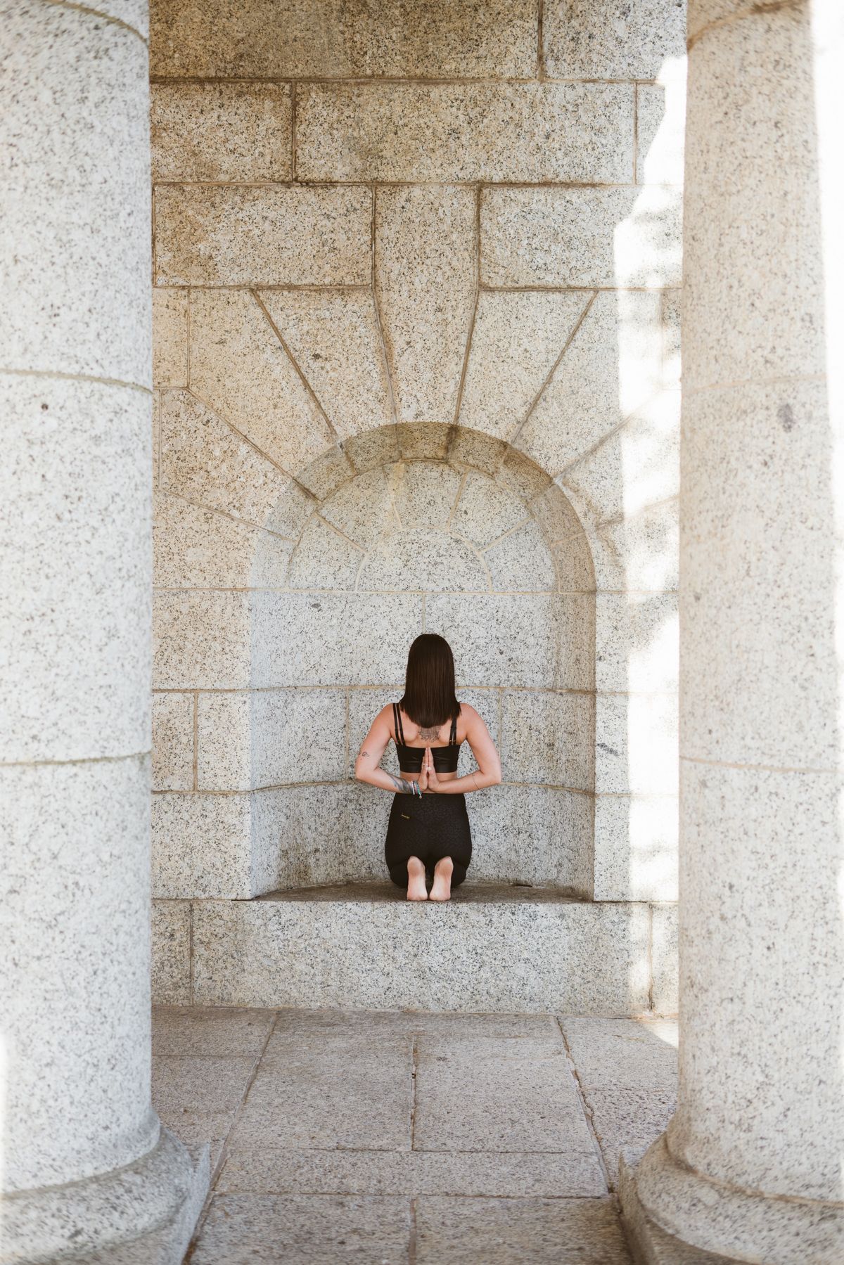 A woman kneels in reverse prayer pose in an alcove surrounded by stone columns.