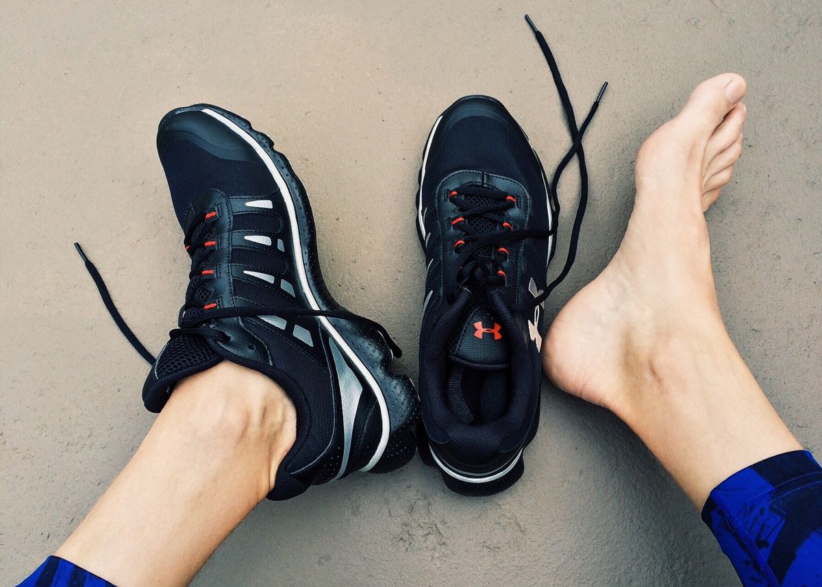 The lower legs of someone wearing one running shoe with a bare foot next to a second running shoe. 